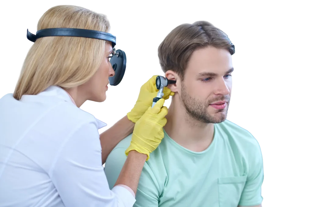 An ENT specialist wearing a head mirror and yellow gloves using an otoscope to examine the ear of a male patient in a green T‑shirt.