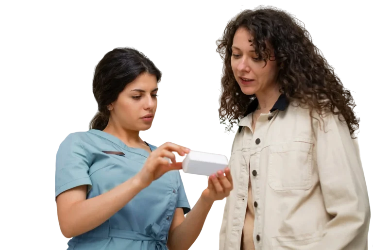 A nurse in teal scrubs showing a medication box to a standing patient in a beige jacket, explaining dosage instructions.