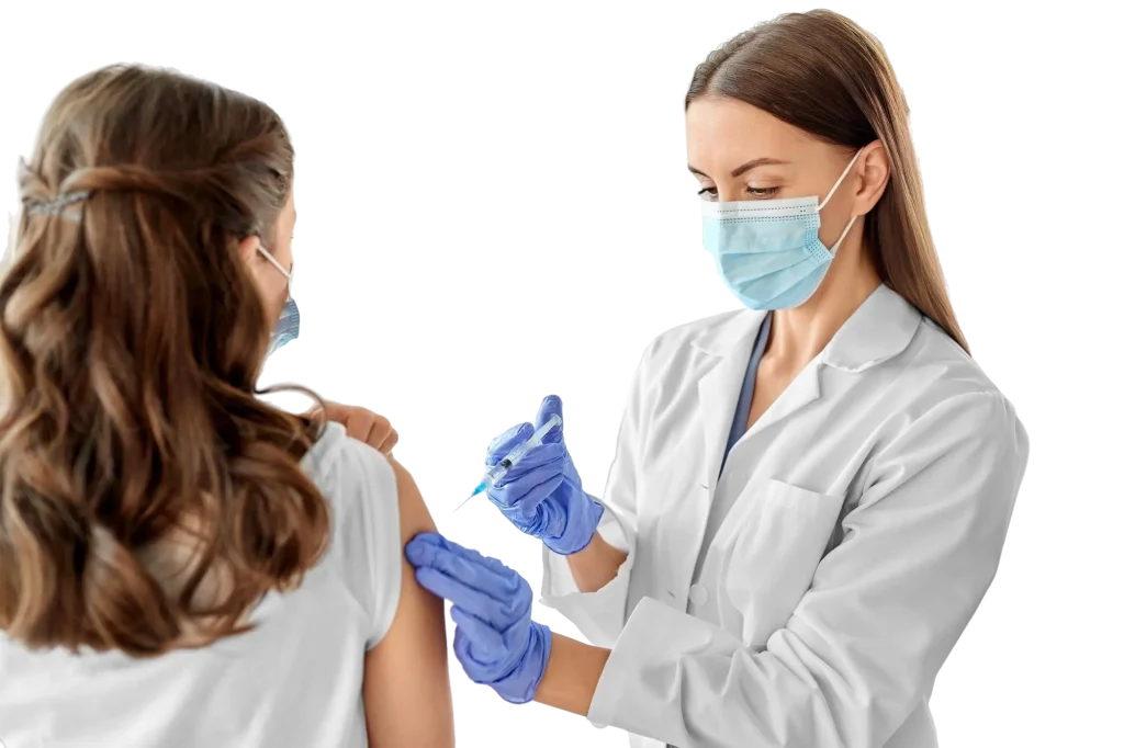 A healthcare professional in a white coat and blue gloves administering a vaccine injection into the upper arm of a seated patient wearing a white T‑shirt.